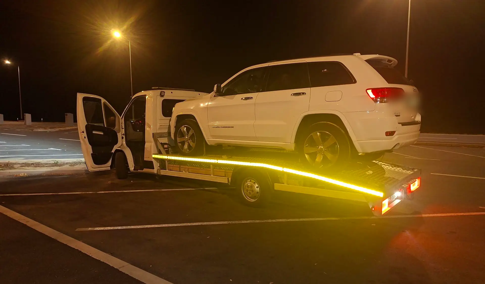 Bijeli kamion AUTO MIRO s osvijetljenom platformom i utovarenim bijelim SUV-om noću na parkiralištu